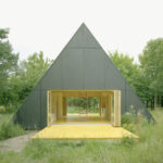 Modernes, dreieckiges Sommerhaus in einer natürlichen Umgebung auf Wolin. Das Haus hat eine auffällige A-Form und ist komplett in einem dunklen, fast schwarzen Ton gehalten, was einen starken Kontrast zum leuchtend gelben Holzdeck davor bildet.