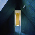 Ein enger Durchgang in einem blau getäfelten A-förmigen Dachboden führt zu einer offenen Tür, durch die man einen Raum in warmen Holztönen und Tageslicht erblicken kann.