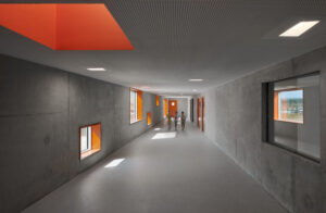 Schulflur in Sichtbeton mit orangefarbenen Akzenten bei Fensterbrüstungen und Oberlicht