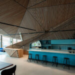 Öffentliches Wohnzimmer mit Kaffeebar und prägnant geformter Holzdecke im Wohngebäude Jonas' von Orange Architects