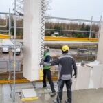Zwei Bauarbeiter setzen einen großen Betonplattenabschnitt mittels eines Krans in eine Gebäudestruktur ein