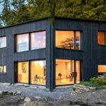 Haus im Grünen mit schwarzer Fassade und hell erleuchteten Fenstern. Reduzierte Architektur mit karbonisiertem Holz.