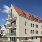Helles Haus mit rotem Dach und Balkonen links: : Nachhaltige Ziegel-Bauweise.