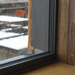 Untere rechte Ecke eines bodentiefen Fensters mit dunklem Rahmen vor braunem Boden und brauner Wand. Draußen weißer Schnee.