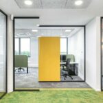 Hellgrüner Boden, an heller Decke zwei Leuchten. Hinten: offene Rahmen mit Trennwandsystem und gelber rechteckiger Farbfläche..