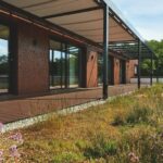 Naturwiese vor rotbrauner Hausfassade mit Fenstern und schmaler Terrasse davor: Holzverbundwerkstoffe.