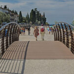 Brücke mit Massivdielen und Geländer. Zwei Personen von hinten gehen auf weiße Promenade mit anderen Menschen zu.